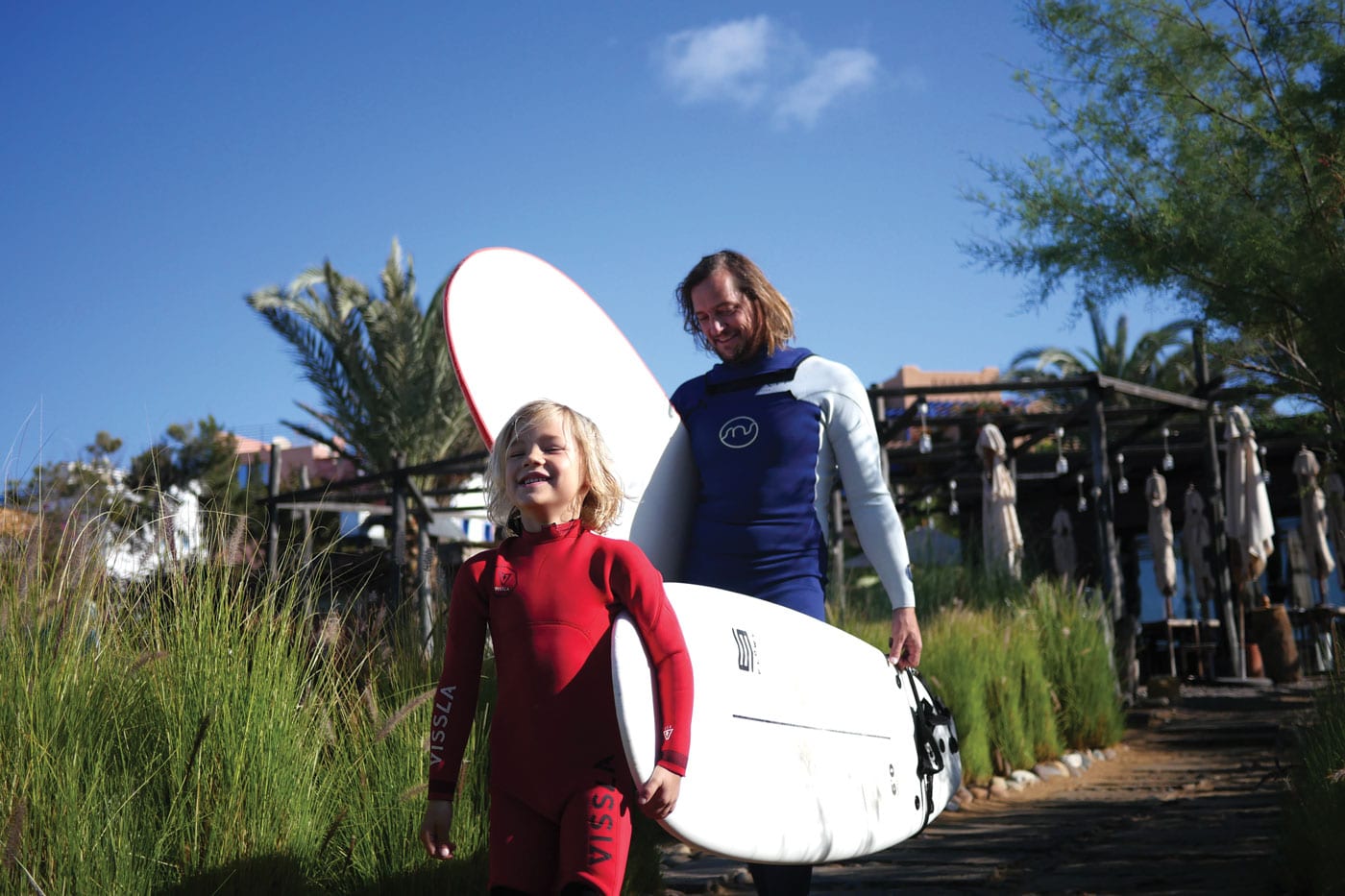A dad and his young son walking with two surf boards in their wetsuits. 